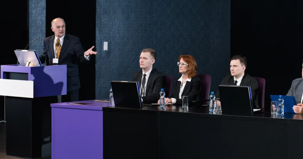 Prelegent przy mównicy i paneliści za stołem prezydialnym podczas międzynarodowej konferencji o zarządzaniu BHP